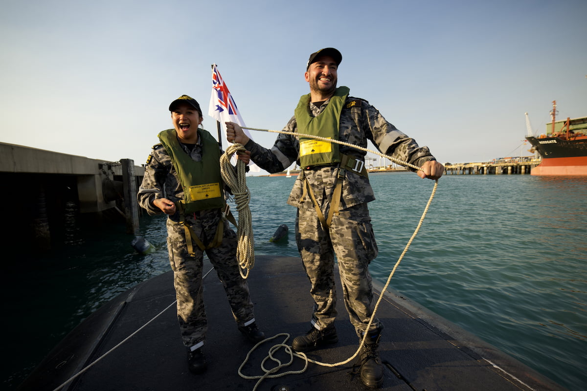 Two people in navy uniform stand on a boat smiling.