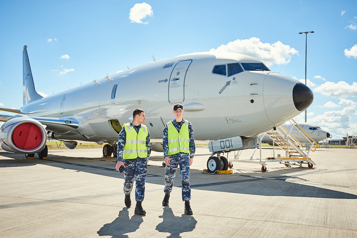 Two young men are on the runway standing in front of a plane.