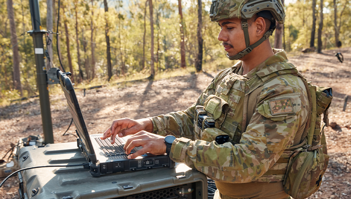 Out in the field, a member of the Army works on his laptop.
