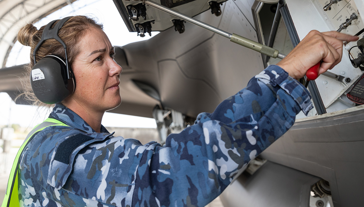 A woman in the Air Force  works on an aircraft with a tool in hand.