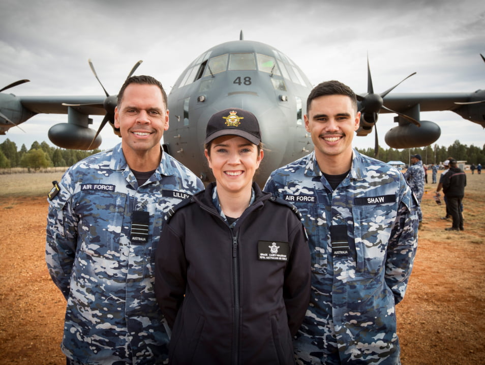Three Air Force members stand outside smiling in front of an airplane.