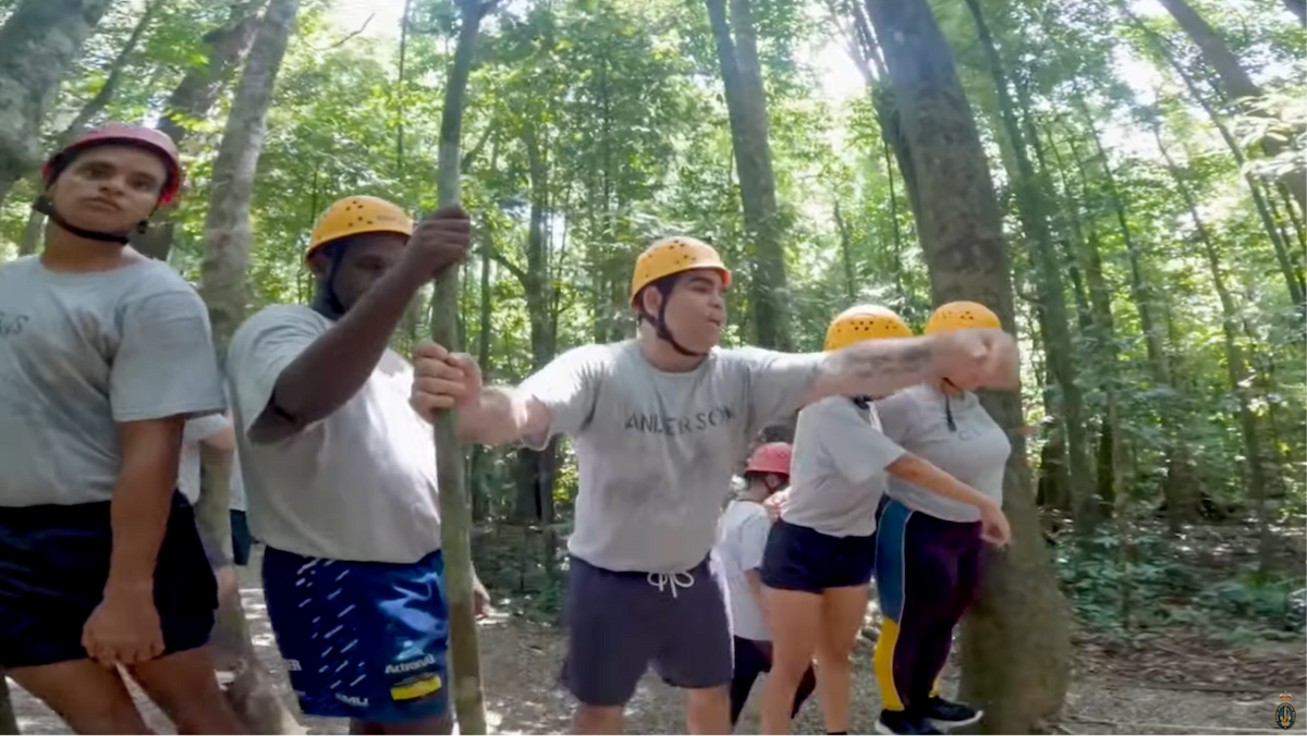 A group of people wearing helmets and harnesses, connected by a rope, engaged in a team-building activity.